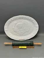 Photo showing the white glazed oval turkey platter with embossed design propped vertically behind the green marble rolling pin with light wooden handles placed horizontally in front.