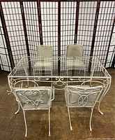 Front view of vintage white wrought iron rectangular glass top patio table with four matching chairs with beige cushions, displayed indoors on wood floor with folding screens in background.