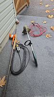 Full shot showing Yardworks grass trimmer with green body, extended shaft and loop handle, orange Black & Decker hedge trimmer, black power cord with remote switch, and orange/red power cord.