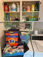 Laundry cabinet with cleaning liquids on shelves, towels stacked below, pails and detergent box on washer and dryer surface.