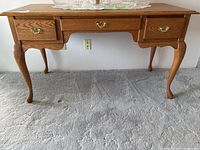 Front view of oak desk showing three drawers, cabriole legs, and brass handles. Surface with decorative lace doilies and glass items.