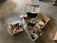 Wide view of lot showing plastic bins filled with paint brushes, rollers, trays, paint cans, and tools on a concrete floor.
