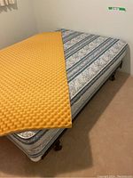 Queen size mattress with blue and white striped pattern, resting on box spring and metal frame. Yellow convoluted foam cushion topper partially covering mattress.