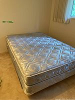 Queen size mattress and box spring set with beige quilted floral pattern in a corner of a carpeted room.