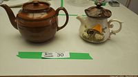 Photo showing both teapots side by side on a table with auction tag 30. Brown Betty teapot is rounded and brown; mushroom teapot is cream with colorful decoration.