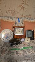 Seven clocks and weather stations displayed on a floral couch, including a large floral wall clock, rectangular wood-framed wall clock, black digital alarm clock, small white bell alarm clock, and small metal clock with star face.
