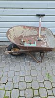 Old rusty metal wheelbarrow with one black pneumatic Numatic tire, viewed from front angle. Inside the wheelbarrow are two metal sprinklers and a Norco tire pump.
