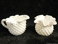 Front view of Fenton hobnail milk glass creamer with handle and hobnail milk glass crimped rim vase on black background