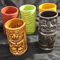 Five tiki mugs of various colors and designs photographed together on black fabric.