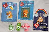 Photo showing five Care Bears figures and magnets on a flat surface, including two magnets in original blue packaging and three small plastic figures, plus one figure attached to a rainbow card.