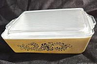 Side view of the Pyrex 502-B Homestead refrigerator dish showing yellow dish body with black floral design and fitted ribbed glass lid