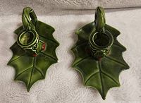 Top view showing both ceramic candle holders shaped as green holly leaves with handles and red berry decoration on a soft fabric background.