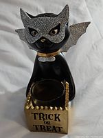 Front view of black ceramic cat candle holder with silver glitter mask and bat wings, gold collar, holding gold candle cup with 'Trick or Treat' text.