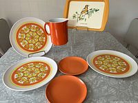 Photo showing three large patterned plates, two smaller solid orange plates, and a small orange pitcher.