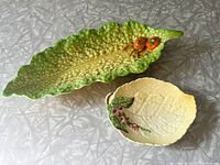 Two Carlton Ware ceramic dishes displayed on a gray textured surface. The larger dish is shaped and colored like a green lettuce leaf with two red tomatoes, the smaller yellow dish resembles a foxglove leaf with floral decoration.