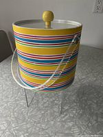 Side view of colourful ice bucket showing multicoloured horizontal stripes and clear plastic handle with yellow knob on lid.