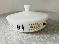 Side view showing white casserole with black diamond and gold harlequin design on the side and matching lid with knob handle.