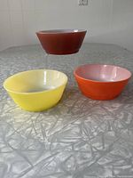 Three vintage Anchor Hocking Fire King bowls in orange, yellow, and dark red arranged on a gray table surface showing exterior colors and shape.