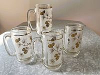 Photo showing four vintage frosted glass beer mugs with gold pinecone and pine needle motifs, arranged on a table.