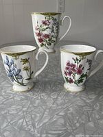 Three McIntosh fine bone china coffee mugs arranged on a table, each with a different detailed floral pattern and gold trim visible on rim and base.