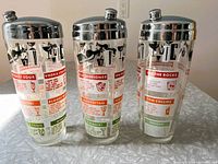 Set of three mid-century modern glass cocktail shakers with chrome tops, shown standing side-by-side with multi-colored recipe print visible around the glass.