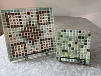 Two mid-century modern square tile dishes displayed on black metal stands on a patterned white surface, showing front tile mosaic pattern and shape.