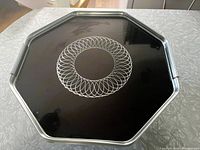 Full top view of octagonal metal bar tray showing black surface with etched white spiral geometric circle design at center and metal rim edge with integrated handle.