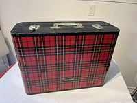 Closed vintage plaid metal suitcase with black top and handle, used as a picnic table. Showing metal clasps and clear handle on top.