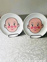 Two white ceramic plates with cartoon face on surface viewed side by side on plate stands