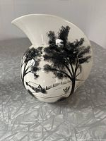 Front side view of white ceramic pitcher with black farm scene depicting trees, barn, fence and field, showing the shape and detail of the handle.