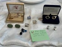 Wide shot of the collection showing cufflinks and tie clips laid on white fabric with a green note describing the lot.