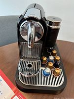Front view of black Nespresso coffee machine with several espresso pods on its tray and milk frother behind.