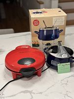 Photo showing red quesadilla maker, blue Chantal fondue pot with stand, and fondue forks beside fondue box.