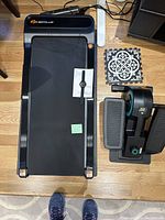 Top view of GoPlus walking pad under desk treadmill with watch and instruction manual on top, placed on wooden floor with rug visible.