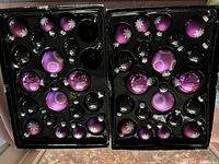 Two black plastic trays holding purple and black glass ornaments, some with snowflake and glitter ring details, total 38 pieces in both trays.