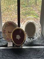 Three picture frames displayed together on a black cloth with measuring tape on table; frames include an oval glass frame with floral embossed design, an oval brass frame with maroon display background, and a cream-colored resin frame decorated with sculpted roses.