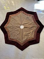 Full view of star-shaped Christmas tree skirt showing burgundy velour border, beige center, embroidered trim, and tree trunk opening.