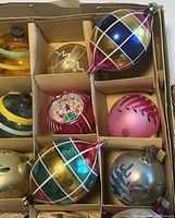 Top-down image showing assortment of large vintage glass Christmas bulbs, some tear drop shaped, with colorful hand-painted and striped designs, housed in sectioned cardboard box.