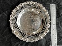 Top view of round silver coaster with ornate grape and vine detailing and pierced lattice rim. A ruler is placed beside for size reference, showing approximately 7 inches diameter.
