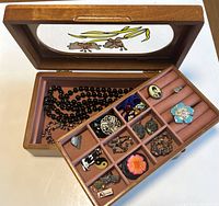 Open wooden jewelry box showing glass top with floral motif and internal tray with various compartments holding pins, rings, and other jewelry pieces.