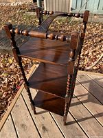 Three-quarter view of wooden shelf showing turned spindle legs, three shelves, and detailed top handle, placed outdoors on wooden surface with fallen leaves in background.