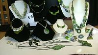 Multiple green costume necklaces displayed on black and white stands, along with earrings and a ring laid out in front, showcasing various bead sizes, shapes, and designs.