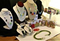 Display of most necklaces on bust stands including multicolored and flower bead designs with bracelets displayed on a stand. Several bracelets and eyeglass holder straps in containers and laid out on the table.