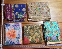 Five folded batik fabrics laid on a wooden surface, colorful floral and butterfly patterns visible.