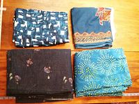 Four folded fabrics on wooden surface, showing variety of patterns and colors: dark batik, blue sun dots, blue and white geometric and figural prints.
