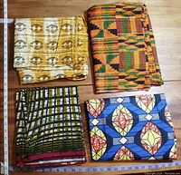 Four folded West African fabrics with different colors and patterns placed on wooden floor alongside rulers for size reference.