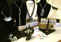 Photo showing 4 necklaces on black bust stands, numerous silver-coloured bangles on display stand, 4 rings on ring holder, and several pairs of earrings arranged on black background.