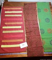 The lot of three fabric pieces lying flat on a wooden table: the orange/red runner with stitched panels on the left, the metallic vine embroidered reddish-brown scarf in middle, and green scarf with circular motifs on right.
