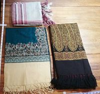 Three shawls laid flat on wooden floor displaying patterns and fringed edges. Top left smaller solid shawl with white and maroon stripes, bottom left greenish-blue and beige with paisley border, right side green and beige paisley pattern on black and purple reversible fabric.