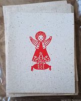 Front of a Christmas card showing a red stylized angel design on textured beige flecked paper.